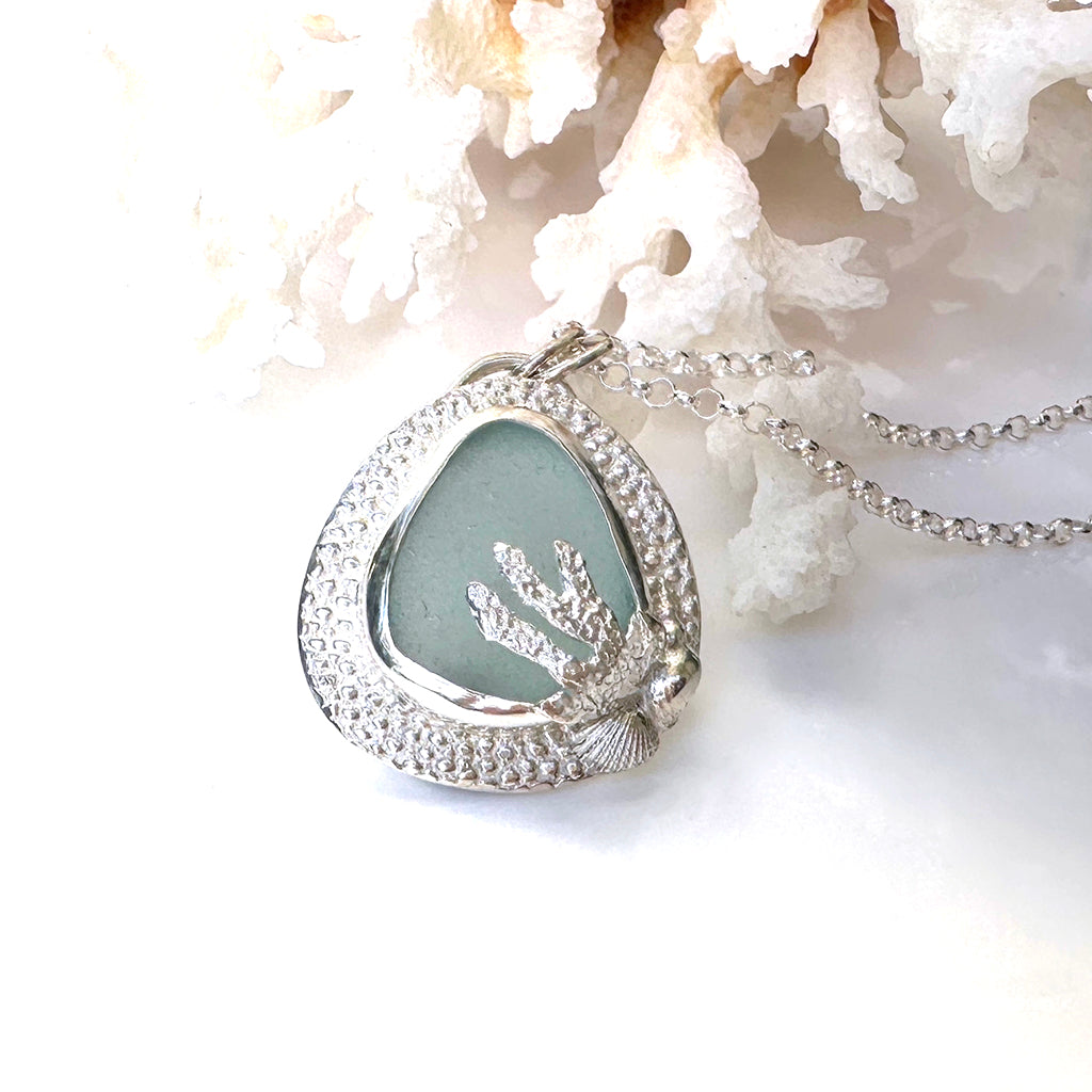 Handcrafted Aqua Mist Sea Glass pendant  bezel set in Manta Ray textured recycled fine silver. Ocean-inspired jewellery by Coastalstyle Australia displayed against neutral background on a piece of coral