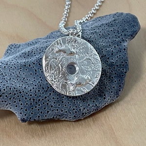Pure silver Sea Urchin Under the Reef Necklace resting on a blue cora-inspired background showing reverse side of 2 little fish swimming amongst the reef