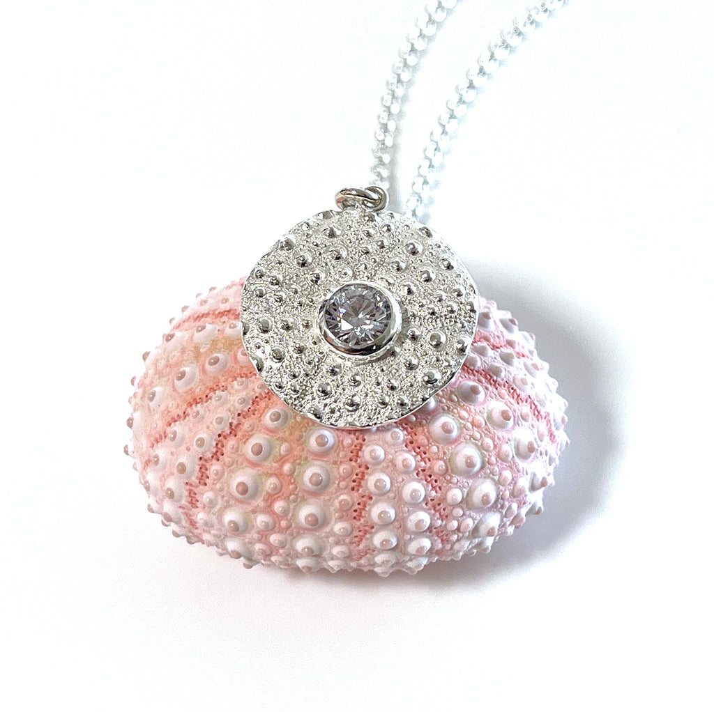 Pure silver Sea Urchin Under the Reef Necklace resting on a seashell-inspired background