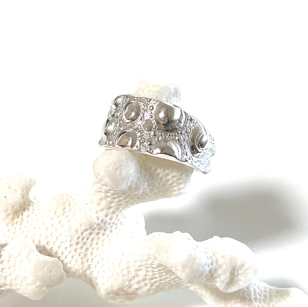 A close-up image of a silver-colored adjustable wide sea urchin ring with a textured surface, displayed on a sea urchin shell.