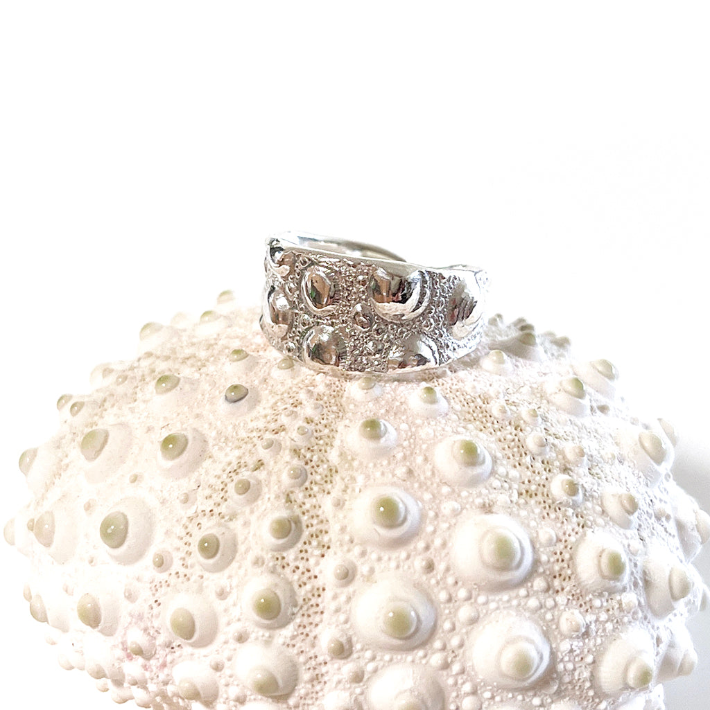 A close-up image of a silver-colored adjustable wide sea urchin ring with a textured surface, displayed on a sea urchin shell.