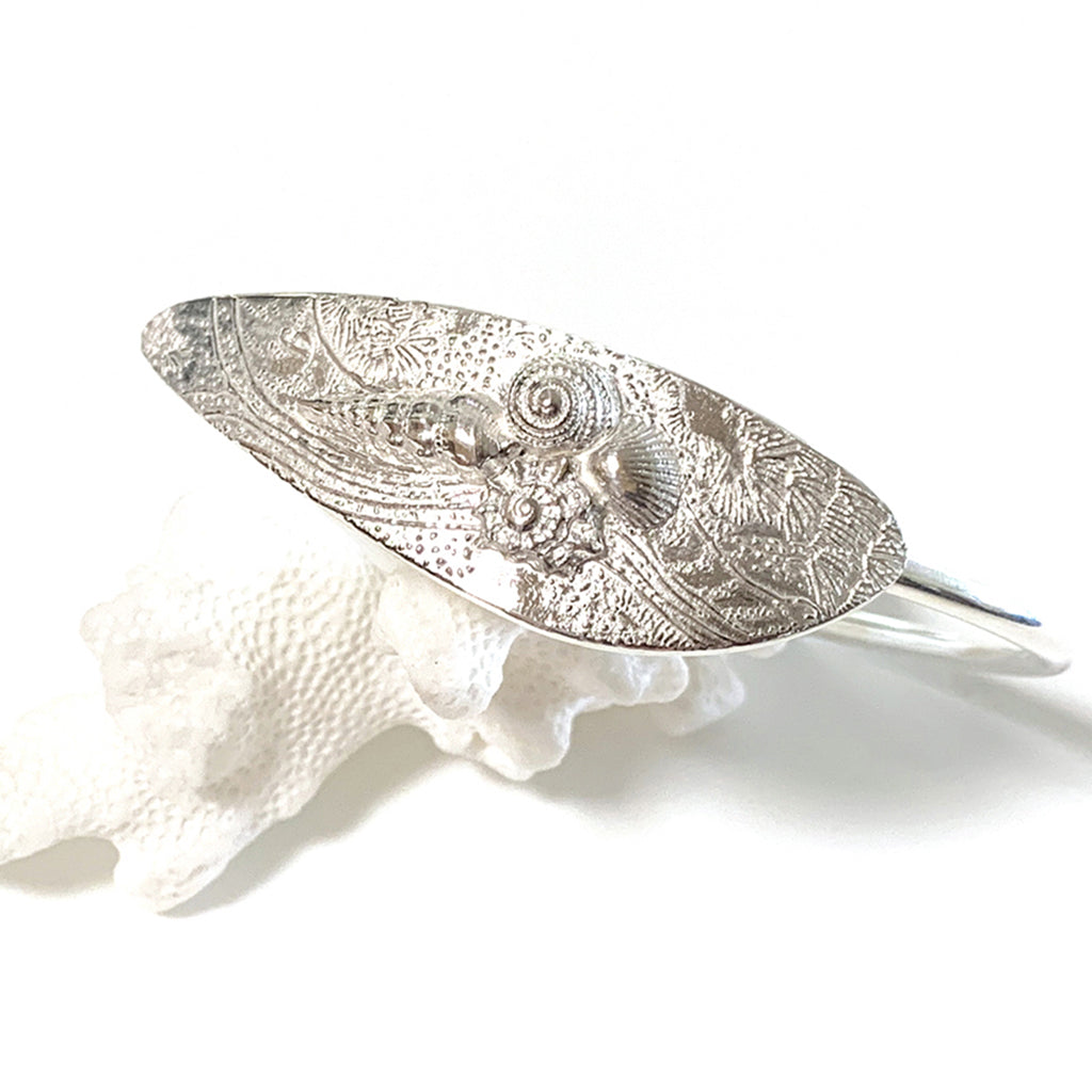 A silver cuff bracelet featuring an ocean-inspired shell design, displayed on a white surface.