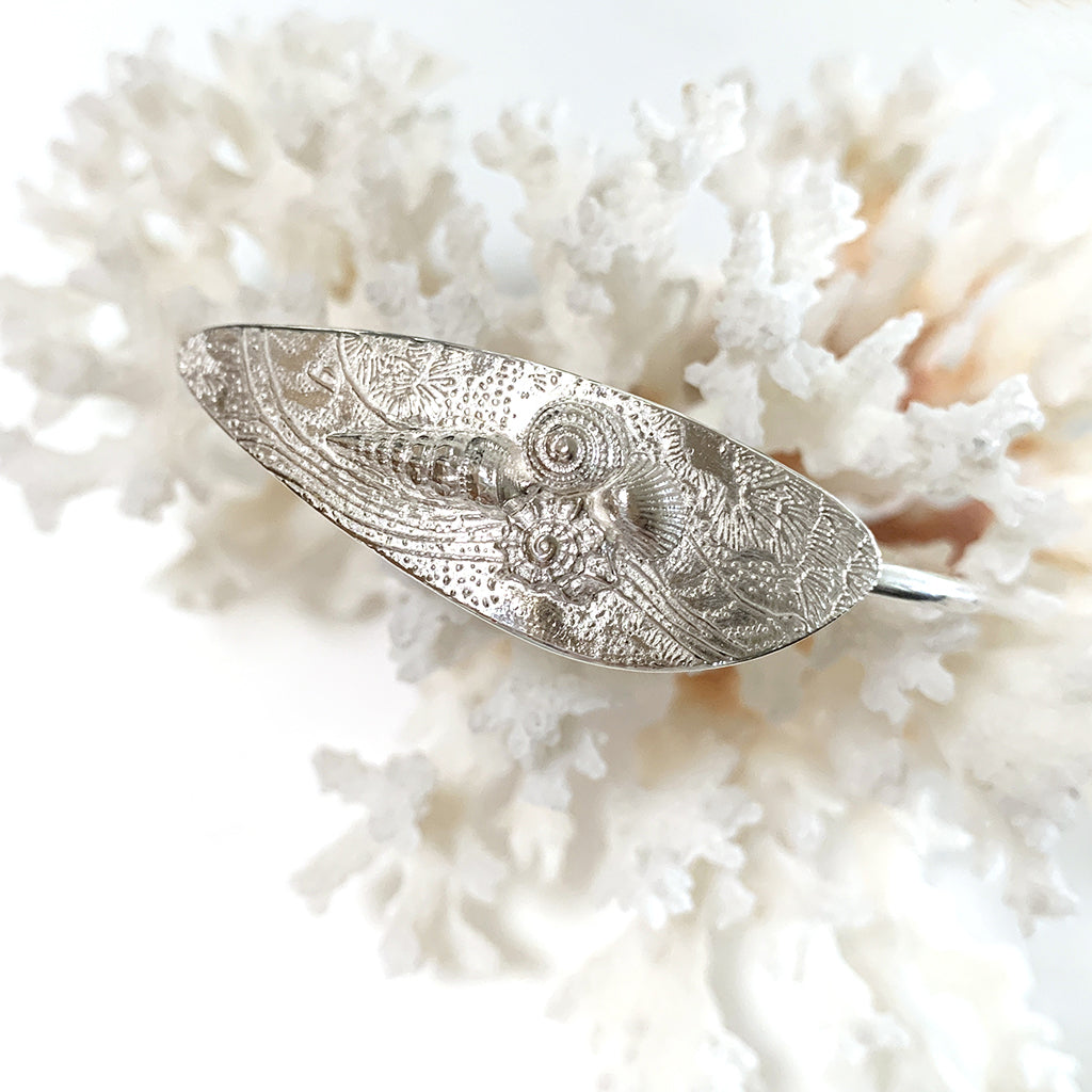 A silver cuff bracelet featuring an ocean-inspired shell design, displayed on a white surface.